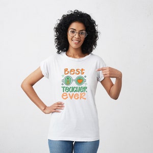 Op de afbeelding: Een wit T-shirt met de tekst "Best Teacher Ever" in oranje en groene letters. De tekst wordt omringd door een afbeelding van een bril met een potlood in het ene glas en een appel in het andere.