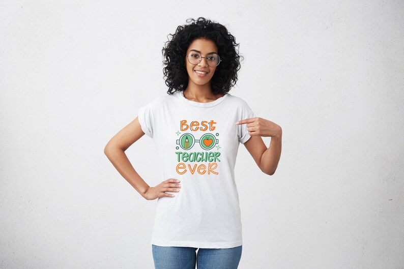 Op de afbeelding: Een wit T-shirt met de tekst "Best Teacher Ever" in oranje en groene letters. De tekst wordt omringd door een afbeelding van een bril met een potlood in het ene glas en een appel in het andere.
