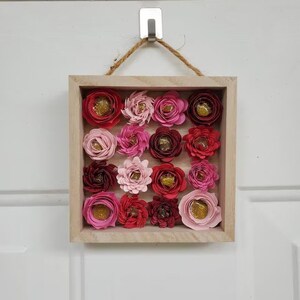 May include: A framed wall decor featuring a collection of 16 paper flowers in shades of pink, red, and burgundy with gold glitter centers. The flowers are arranged in a grid pattern within a light wood frame.