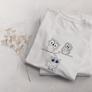 Op de afbeelding: Wit T-shirt met een zwart-witte illustratie van drie uilen op een tak. De uilen hebben grote ogen en kijken in verschillende richtingen. Het shirt is gevouwen en ligt op een wit oppervlak.