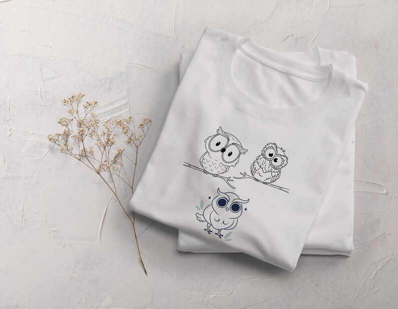 Op de afbeelding: Wit T-shirt met een zwart-witte illustratie van drie uilen op een tak. De uilen hebben grote ogen en kijken in verschillende richtingen. Het shirt is gevouwen en ligt op een wit oppervlak.