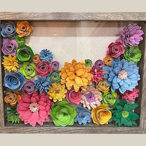 May include: A framed shadow box art piece featuring colourful paper flowers in shades of blue, green, pink, yellow, and orange. The flowers are arranged in a cascading pattern and are set against a neutral linen background.