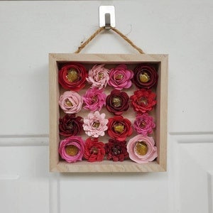 May include: A square wooden frame with a hessian rope hanger, containing a wall art piece made of 16 paper flowers in shades of pink, red, and burgundy. The flowers have a layered, textured appearance and some have gold accents.