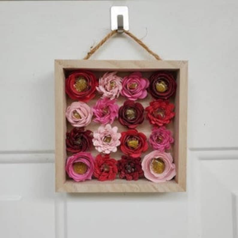 May include: A square wooden frame with a hessian rope hanger, containing a wall art piece made of 16 paper flowers in shades of pink, red, and burgundy. The flowers have a layered, textured appearance and some have gold accents.