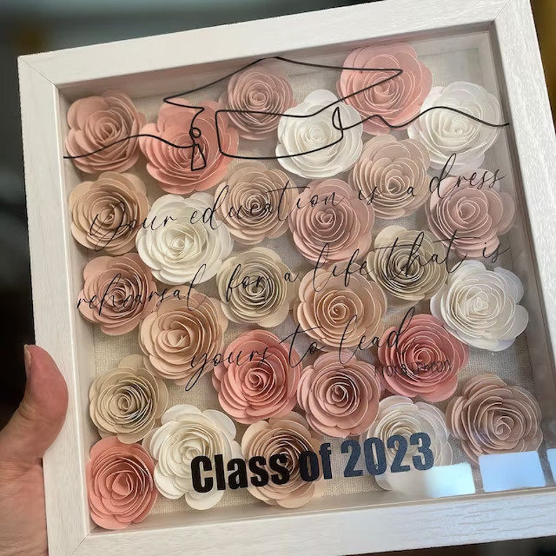 May include: A white shadow box with a glass front filled with pink and white paper flowers. The text "Your education is a dress rehearsal for a life that is yours to lead" is written in black cursive script. The bottom of the box has the text "Class of 2023" in black.
