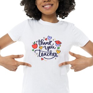 Op de afbeelding: Wit T-shirt met een kleurrijke afbeelding met de tekst "Bedankt leraar" omringd door harten, sterren en een appel.