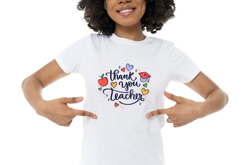 Op de afbeelding: Wit T-shirt met een kleurrijke afbeelding met de tekst "Bedankt leraar" omringd door harten, sterren en een appel.