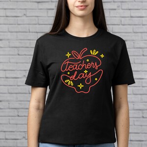 Op de afbeelding: Zwart T-shirt met een rode appel afbeelding waarop "Teachers Day" in een gele omtrek staat.