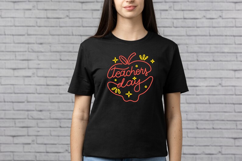 Op de afbeelding: Zwart T-shirt met een rode appel afbeelding waarop "Teachers Day" in een gele omtrek staat.