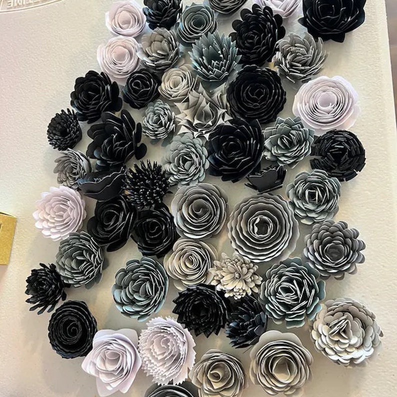 May include: A wall adorned with an assortment of paper flowers in shades of grey and black. The flowers are arranged in a haphazard pattern, creating a visually appealing and textured display.