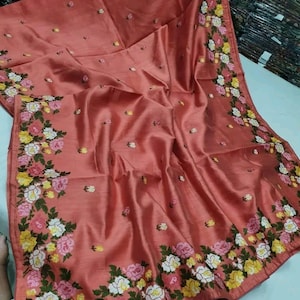 Pode incluir: Um sari de seda cor de pêssego com um design de bordado floral. O bordado apresenta flores brancas, rosas e amarelas com folhas verdes.