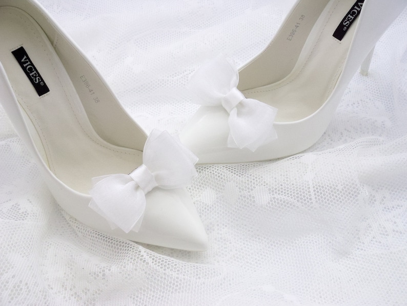 May include: A pair of white satin high-heeled shoes with white satin bows on the toes. The shoes are on a white lace background.