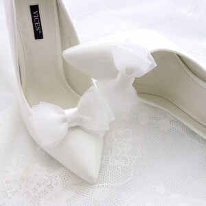 May include: A pair of white satin bridal shoes with a pointed toe and a high heel. The shoes have a white satin bow on the front and the brand name "VICES" is visible on the inside of the shoe.