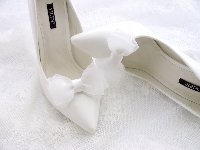 May include: A pair of white satin bridal shoes with a pointed toe and a high heel. The shoes have a white satin bow on the front and the brand name "VICES" is visible on the inside of the shoe.