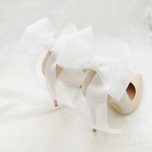 May include: A pair of nude high-heeled shoes with large white satin bows tied around the ankles.