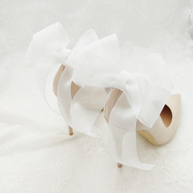 May include: A pair of nude high-heeled shoes with large white satin bows tied around the ankles.