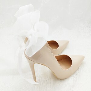 May include: A pair of nude patent leather high heels with a white satin bow on the back of one shoe.