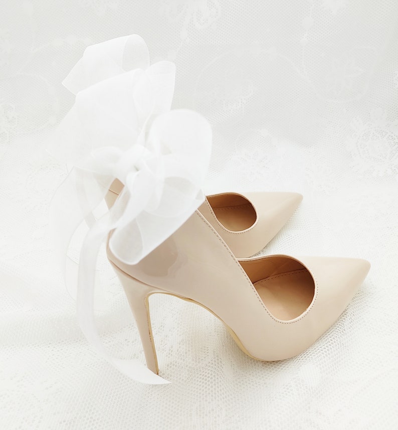 May include: A pair of nude patent leather high heels with a white satin bow on the back of one shoe.