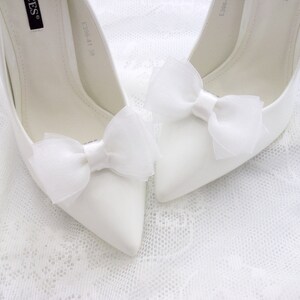 May include: A pair of white satin high heel shoes with white tulle bows on the toes. The shoes are on a white lace background.