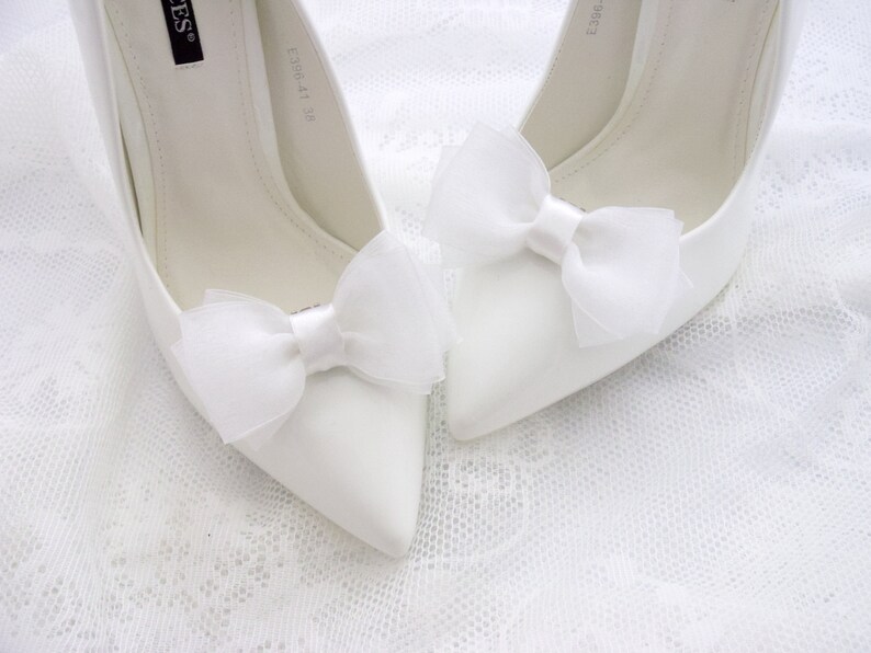 May include: A pair of white satin high heel shoes with white tulle bows on the toes. The shoes are on a white lace background.