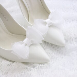 May include: A pair of white satin high heel shoes with white tulle bows on the toes. The shoes are on a white lace background.