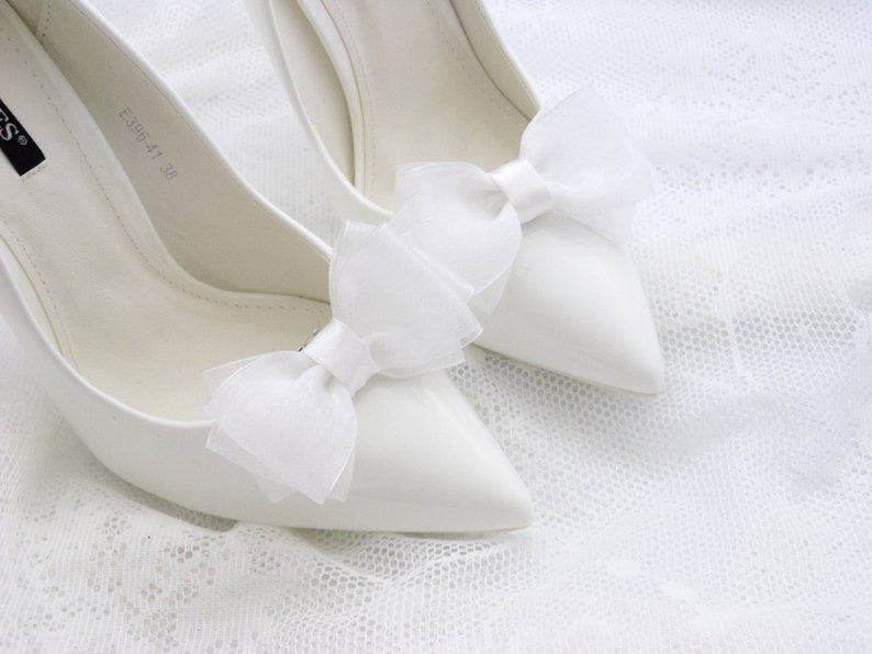 May include: A pair of white satin high heel shoes with white tulle bows on the toes. The shoes are on a white lace background.