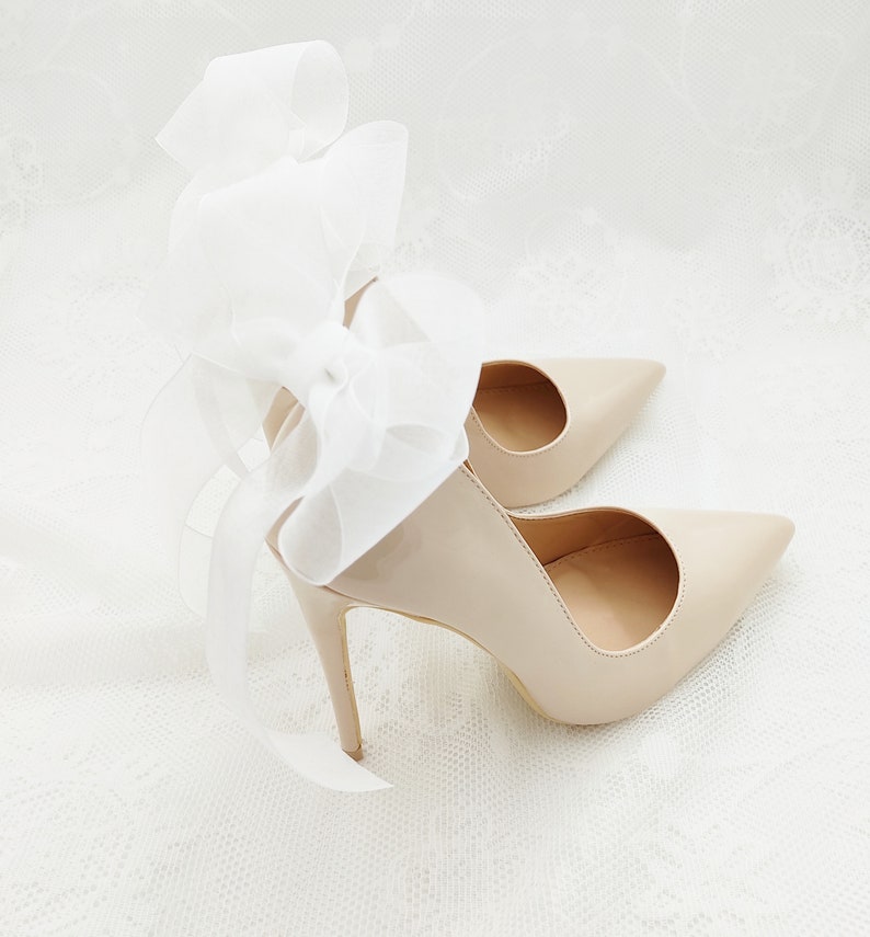 May include: A pair of nude-colored high-heeled shoes with a white satin bow on the back.
