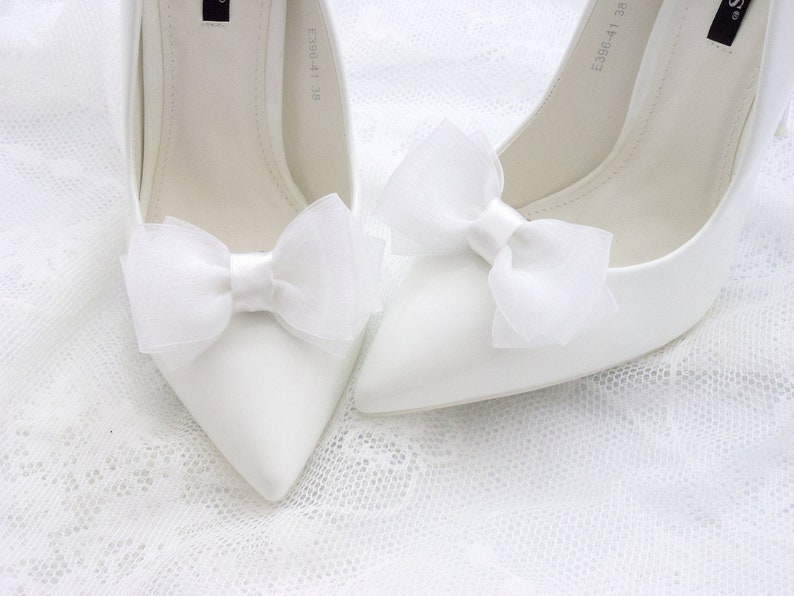 May include: A pair of white satin high heel shoes with white tulle bows on the toes.