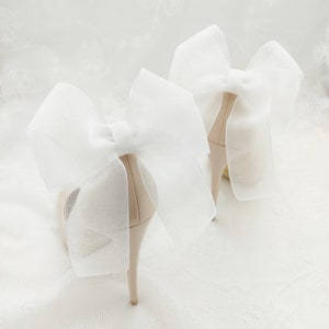 May include: A pair of nude high-heeled shoes with large white tulle bows on the back.
