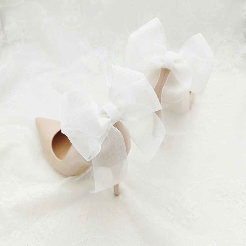 May include: A pair of beige high-heeled shoes with large white tulle bows on the back.