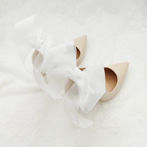 May include: A pair of nude-colored high heels with a white bow on each shoe.