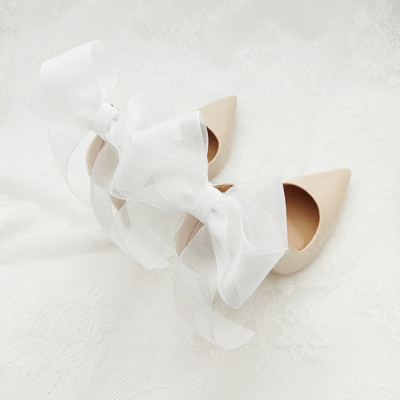May include: A pair of nude-colored high heels with a white bow on each shoe.