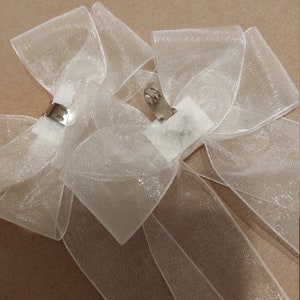 May include: Two white sheer ribbon bows with silver clips. The bows are tied with a simple knot and have a shimmery finish.