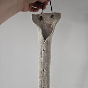 Puede incluir: Un jarrón de pared de cerámica blanco roto hecho a mano con una superficie texturizada y una forma alargada única. El jarrón presenta un diseño floral decorativo y tres pequeños agujeros. Se adjunta una cuerda negra para colgarlo.