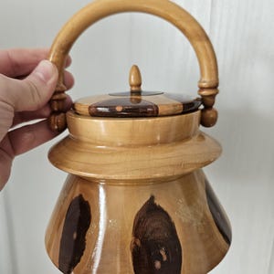 Pode incluir: Um recipiente de madeira artesanal com tampa e alça. O recipiente é marrom claro com padrões de grão de madeira marrom escuro. A tampa e a alça são de um tom mais claro de madeira.