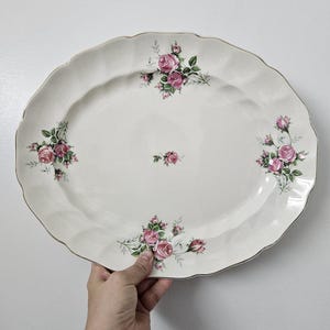 May include: An oval, cream-coloured ceramic serving platter with a scalloped edge and gold trim. The platter is decorated with clusters of pink roses and green leaves. A hand is holding the platter.
