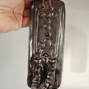 May include: A dark brown ceramic wall hanging with a leather cord. The rectangular piece has raised circular and rectangular patterns, and two decorative, stylised feet at the base. The piece is likely for decorative purposes.