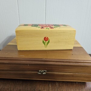 May include: A rectangular light-coloured wooden box with a hand-painted floral design on the lid and a single red flower on the front, resting on a larger, flat, dark-brown wooden box featuring a metal latch closure on the front edge.