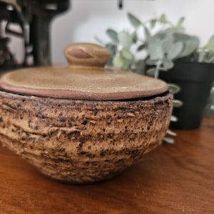 May include: A handmade ceramic jar with a lid. The jar is a mottled brown and tan colour with a textured surface. The lid has a small knob and a darker brown rim. The jar is on a wooden surface.