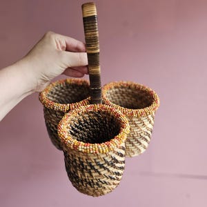 May include: A woven basket carrier featuring three cylindrical compartments joined together. The basket is crafted from light and dark brown natural fibres with a vertical handle. Each rim is decorated with a band of small orange and yellow beads.