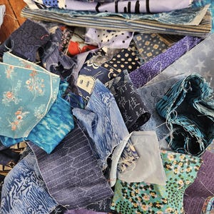 Peut inclure: Une collection de chutes de tissu assorties dans diverses nuances de bleu, présentant des motifs variés incluant des fleurs, des pois, des rayures et des formes géométriques. L'amas se compose de multiples pièces textiles aux bords effilochés.
