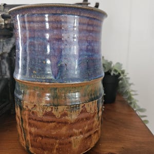 May include: A handmade ceramic jar with a two-tone glaze. The upper part has a gradient of blues and purples, while the lower part shows earthy browns and tans. The jar has a wide opening and a textured surface, suitable for kitchen or home decor.