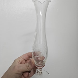 Könnte beinhalten: Eine klare Glasvase mit einem gewellten Rand und einem zarten, geätzten Design. Die Vase hat einen schlanken Hals und einen runden Boden.