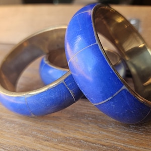 May include: Two circular bangles featuring a polished gold-tone metal interior and a vibrant blue exterior surface. The blue sections are segmented by thin, light-coloured lines, creating a grid-like pattern around the circumference of each bracelet.