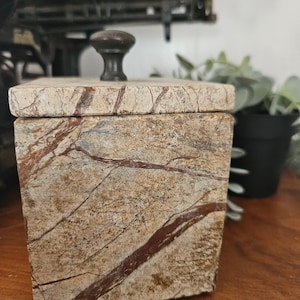 May include: A square stone box with a lid and a small, dark knob. The box is a mix of beige, brown, and grey with natural veining. The lid sits flush on the box, and the knob is centered on top.