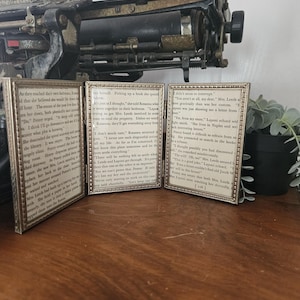 Op de afbeelding: Drie kleine rechthoekige ingelijste prenten met goudkleurige frames. De prenten tonen verouderde pagina's uit boeken met tekst. De frames zijn gerangschikt op een houten oppervlak, met een antieke typemachine op de achtergrond. Vintage decor.