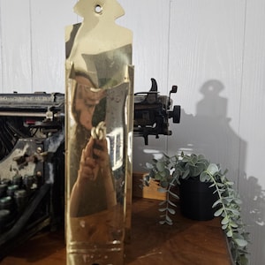 May include: A brass wall pocket, rectangular with a decorative top. The surface is reflective. The background includes a typewriter, a small potted plant, and a wooden surface. The pocket is likely for storing small items.