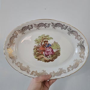 Puede incluir: Fuente ovalada para servir con base color crema y borde de filigrana dorada. El centro presenta una escena pastoral con figuras con vestimenta de época. El borde tiene un borde festoneado.