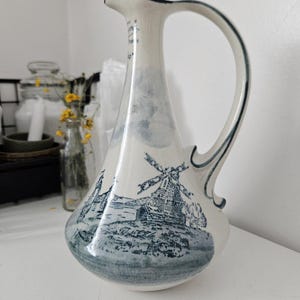 May include: White ceramic jug with a blue and white windmill design. The jug has a curved handle and a spout with a dark blue trim. The design features a landscape with windmills and water. A decorative piece for the home.