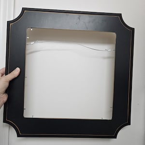 May include: A black picture frame with a decorative, scalloped edge and a thin gold trim. The frame is square with a rectangular opening and a wire for hanging. The frame is held by a hand.
